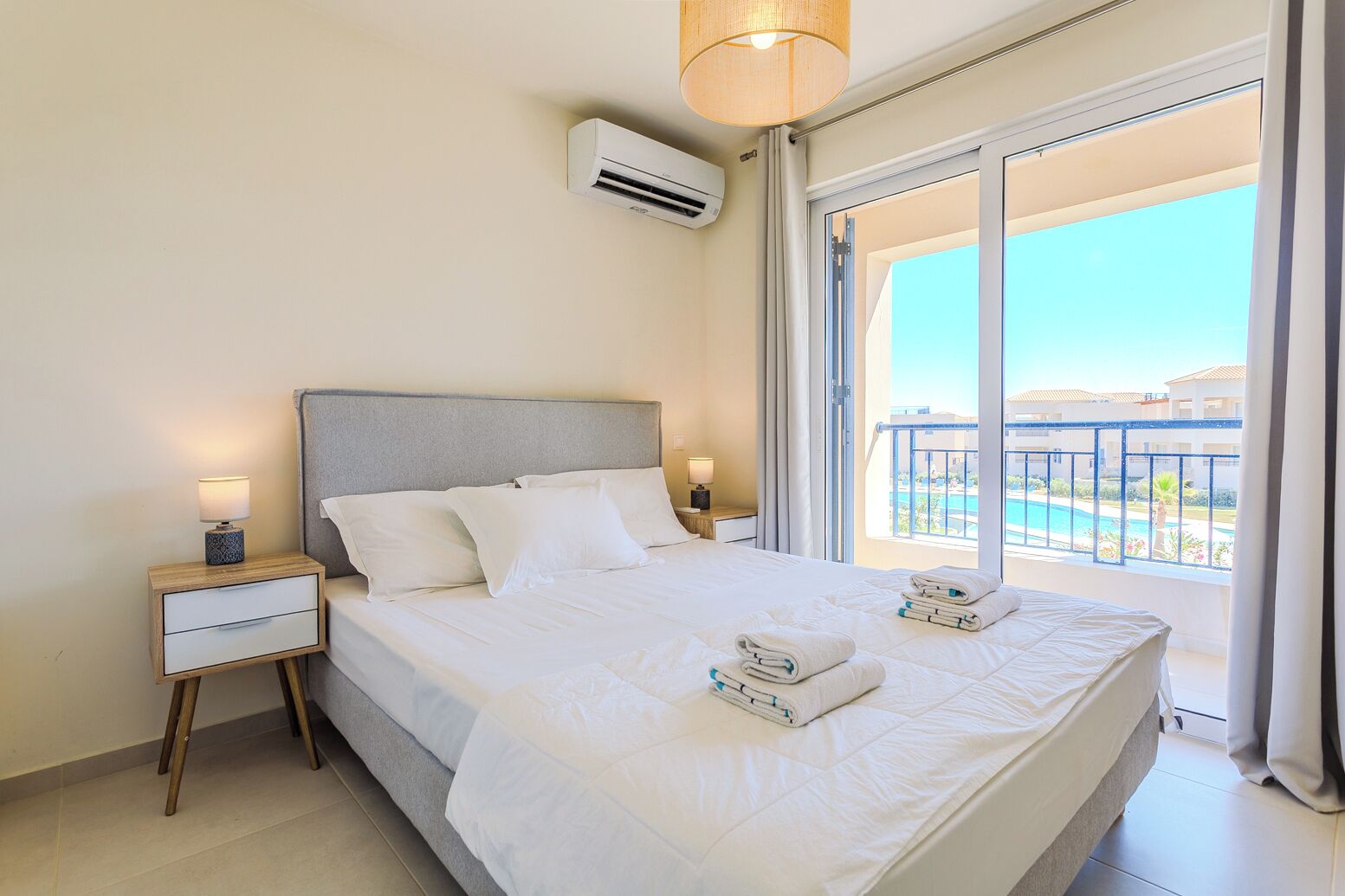Habitación de hotel con cama, mesitas de noche, unidad de aire acondicionado y balcón con vista a la piscina.
