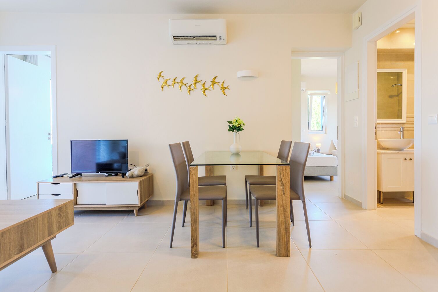 A modern living room with a flat-screen TV, a dining table with chairs, and a decorative wall piece.