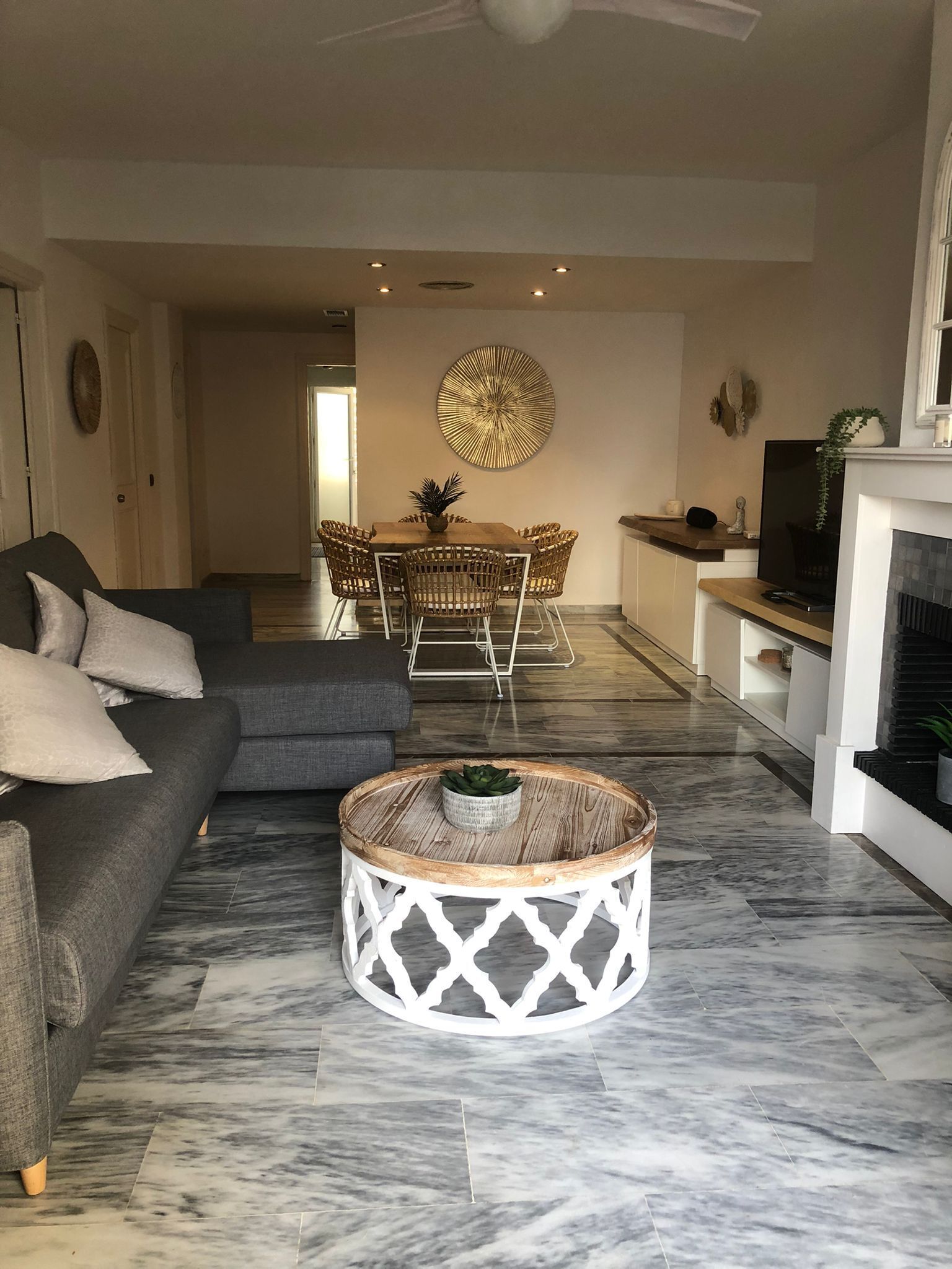 Una sala moderna con un sofá gris, una mesa de centro redonda y un comedor con sillas de mimbre y una mesa central.