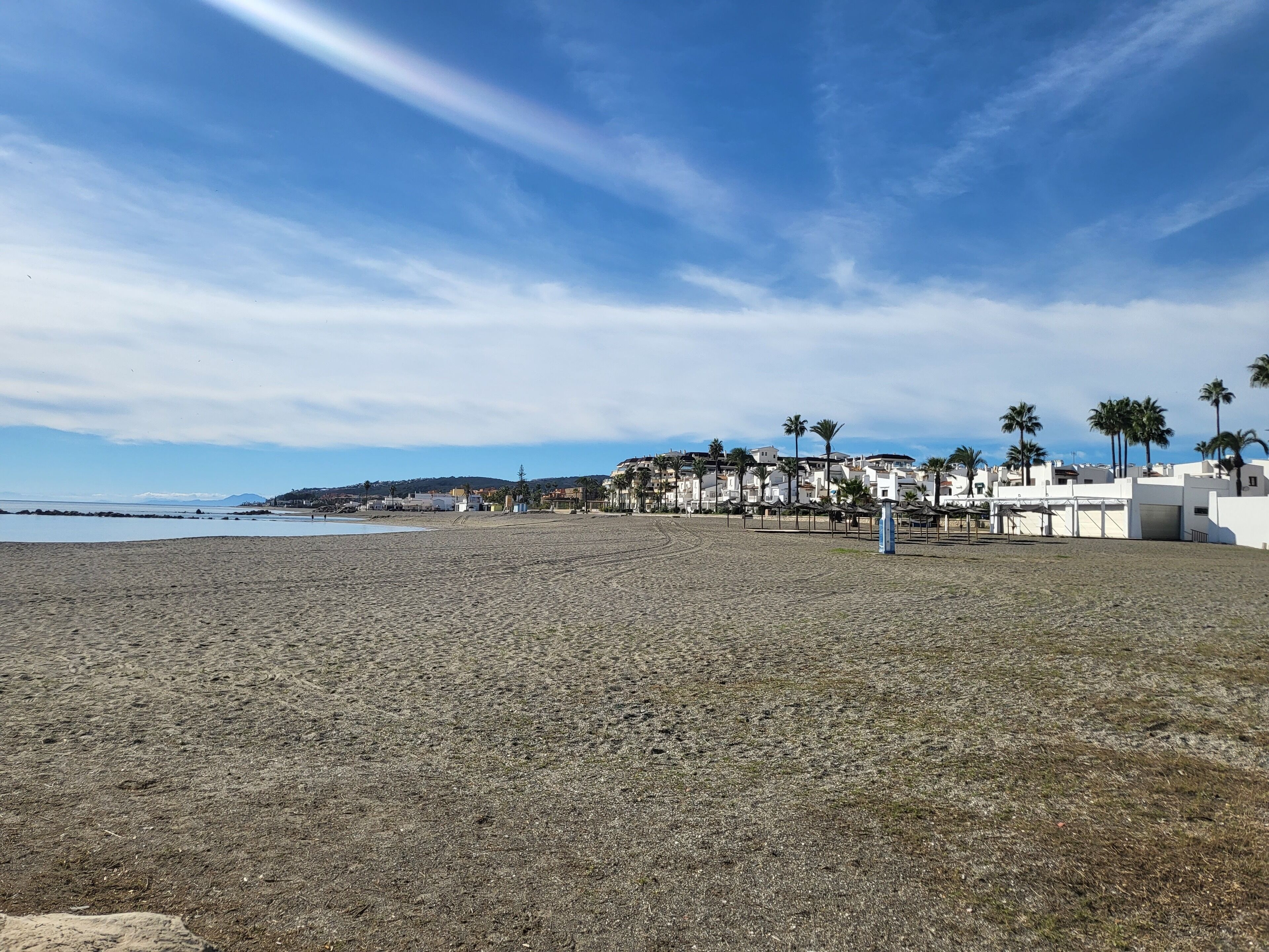 A beach with pebbles, white buildings, and palm trees.
