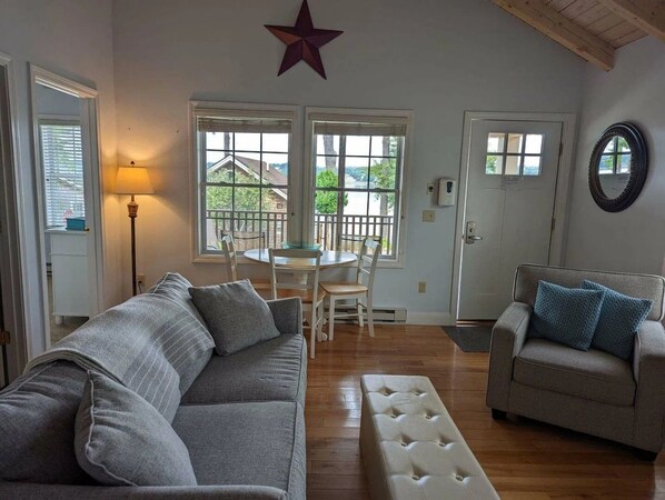 A living room with a sofa, a chair, a bench, and a dining table with chairs. There are windows with blinds, a star decoration, and a door with a mirror. - Waterfront 2BR cottage Sheepscot Harbour Cottage #19 (Edgecomb)