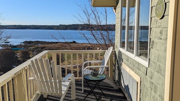 Ett trädäck med två vita Adirondack-stolar och ett litet bord, med utsikt över en vattenyta.