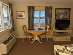A room with a dining table, two chairs, a television on a stand, a heater, and a view of the sea through large windows. - Waterfront studio with kitchen & Wi-Fi Sheepscot Harbour Studio #210 (Edgecomb)