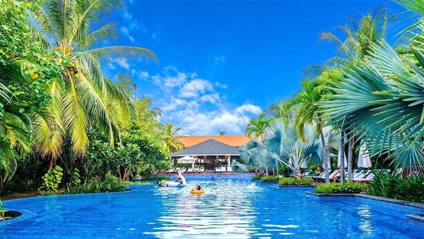 A swimming pool with palm trees, lounge chairs, and a building in the background. - Luxury Pool Villas in Purama Villas (Da Nang)