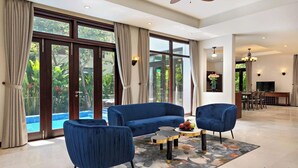 A spacious living area with a blue sofa, two armchairs, and a coffee table. Large windows offer a view of a swimming pool and greenery. - Luxury Pool Villas in Purama Villas (Da Nang)