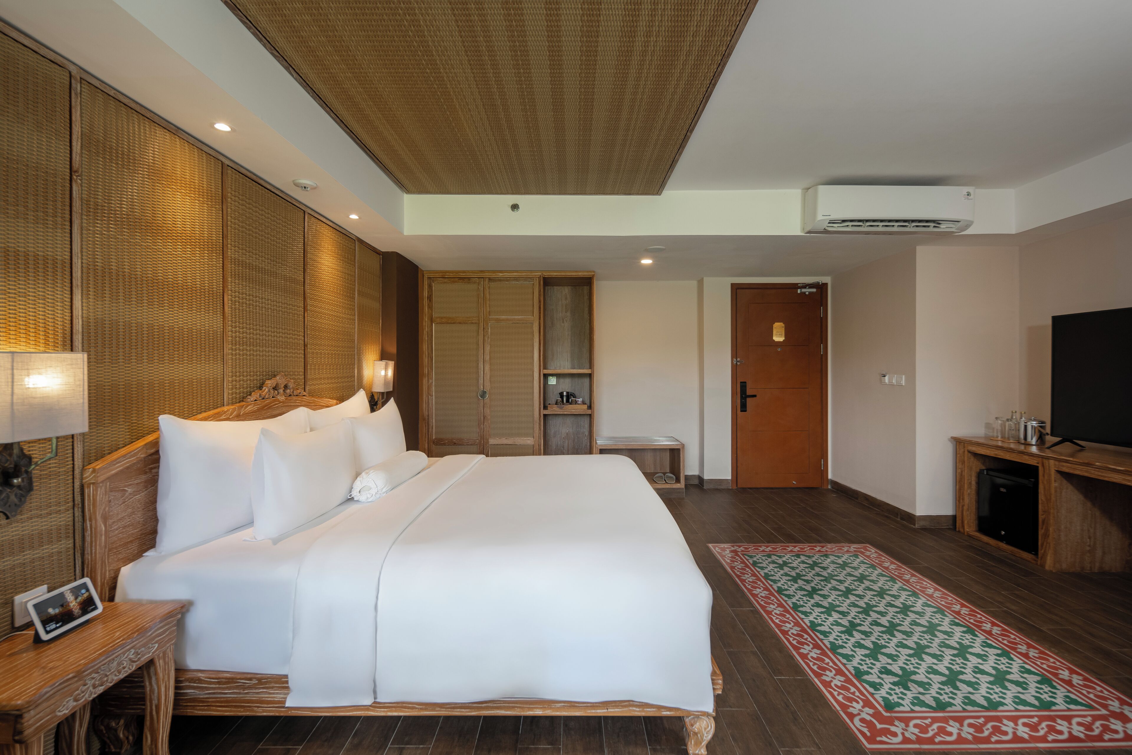 Habitación de hotel con una cama grande, una mesita de noche con lámpara, un armario, un televisor y una pequeña mesa con un marco de fotos.