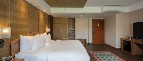 Habitación de hotel con una cama grande, una mesita de noche con lámpara, un armario, un televisor y una pequeña mesa con un marco de fotos.