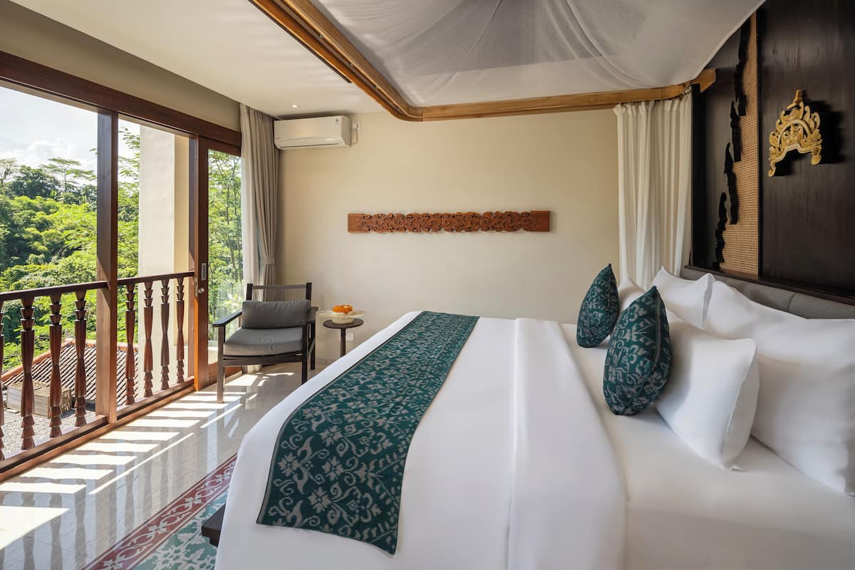 A hotel room with a large bed, a balcony with a view of greenery, a small table with a chair, and a decorative wall piece.