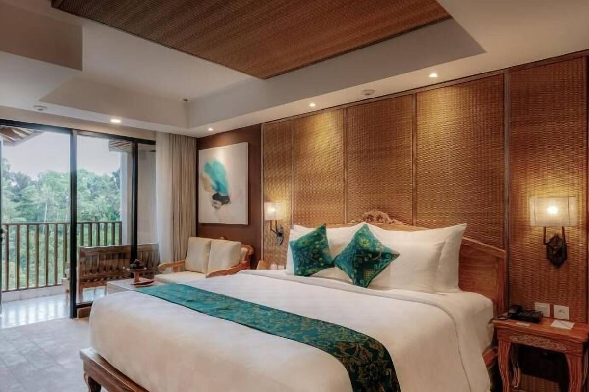 A bedroom with a large bed, a nightstand, a wall-mounted lamp, and a view of the outdoors.