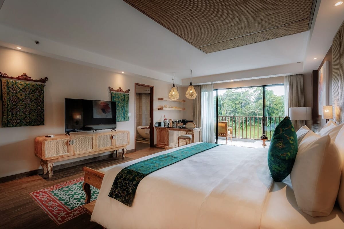 A modern hotel room with a large bed, a television, a dining area, and a balcony with a view.