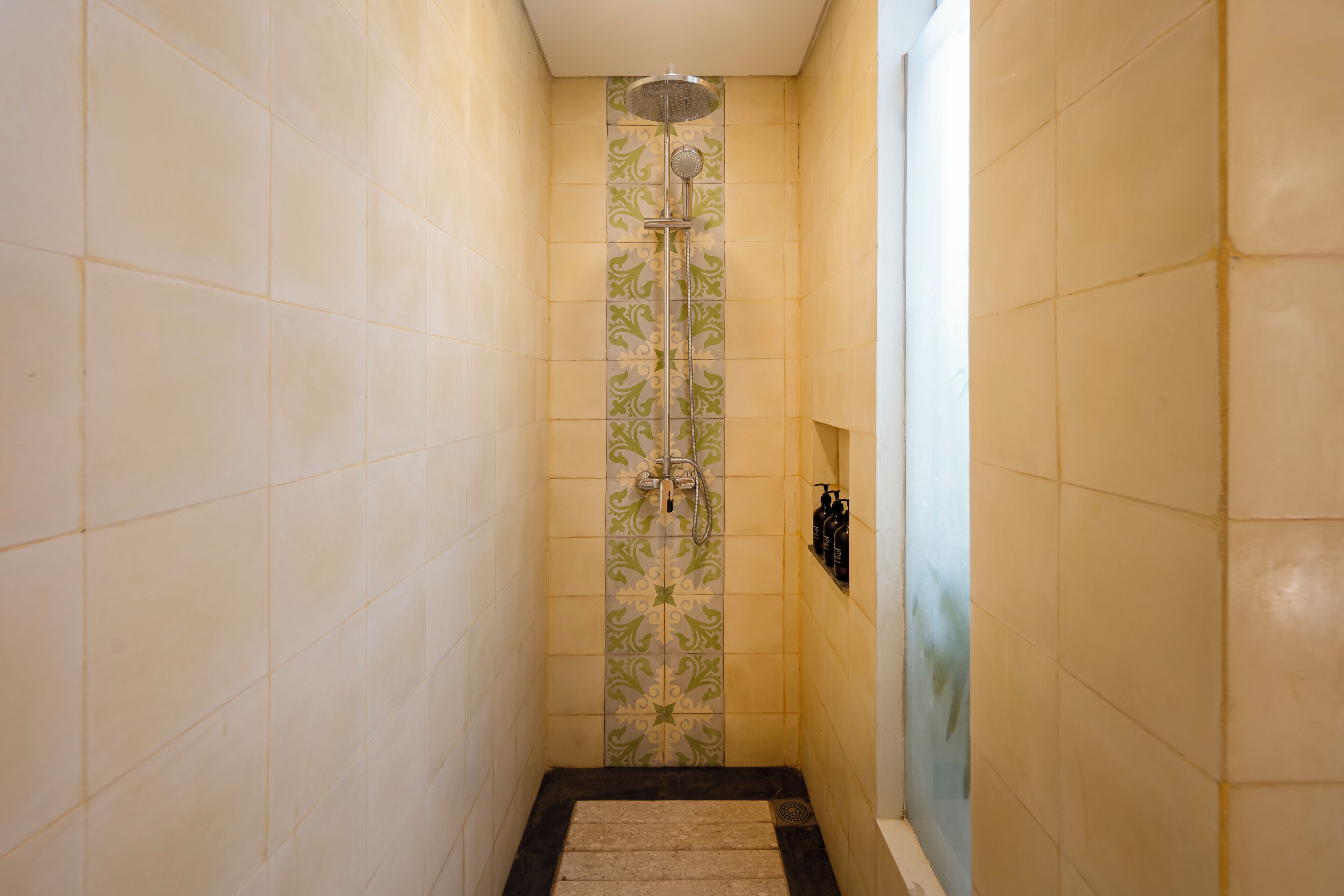 Un espacio de ducha estrecho con una franja vertical de azulejos decorativos, un cabezal de ducha y un pequeño estante con artículos de aseo.