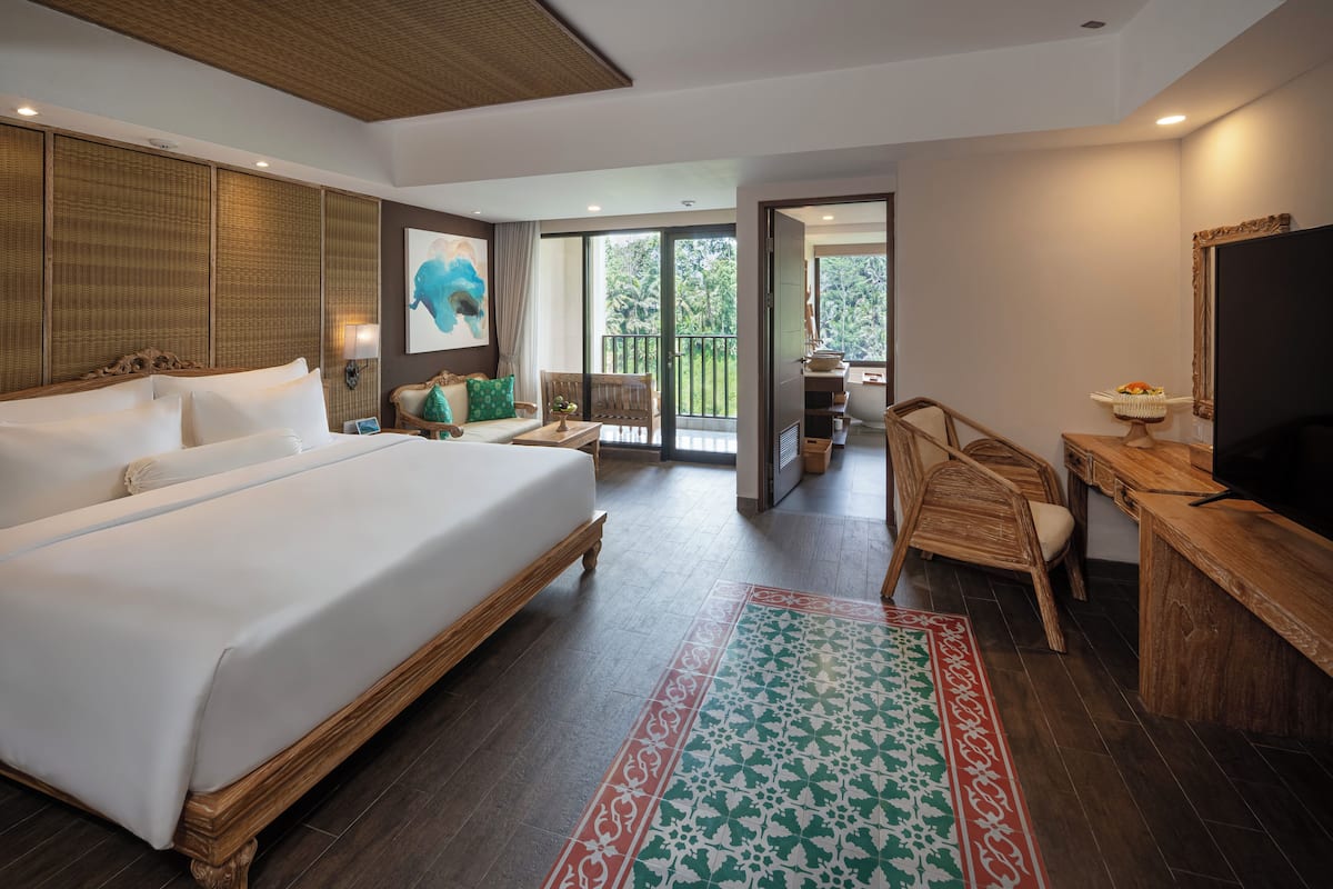 A spacious hotel room with a large bed, a desk, a television, and a balcony with a view of greenery.