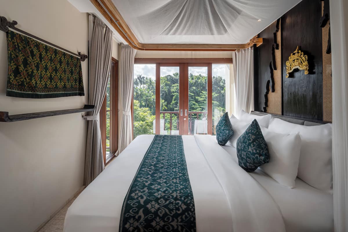 A hotel room with a large bed, a view of lush greenery through the balcony, and a traditional decor style.