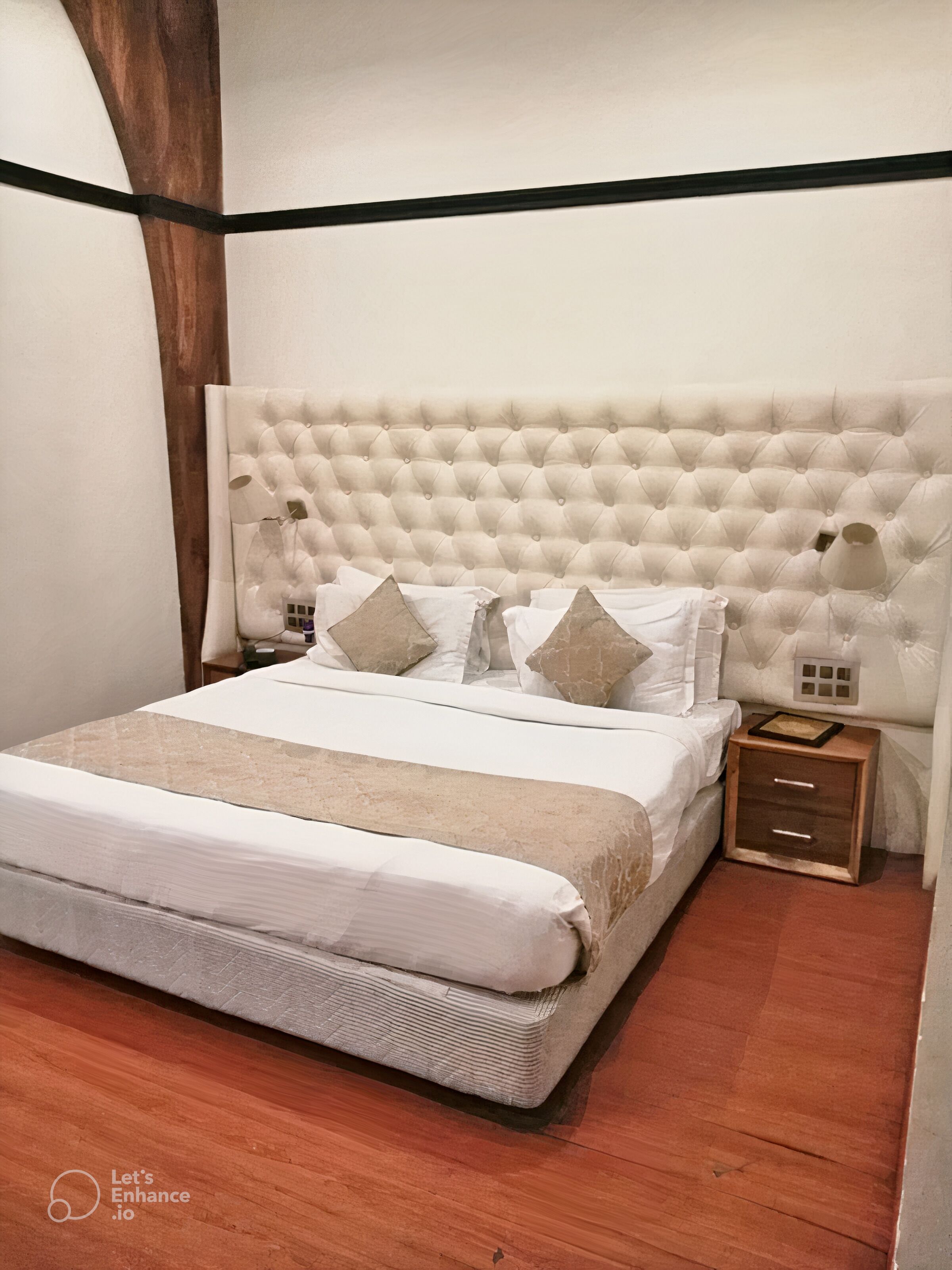 A bedroom with a tufted headboard, a wooden nightstand with drawers, a bed with white bedding, and a wooden floor.