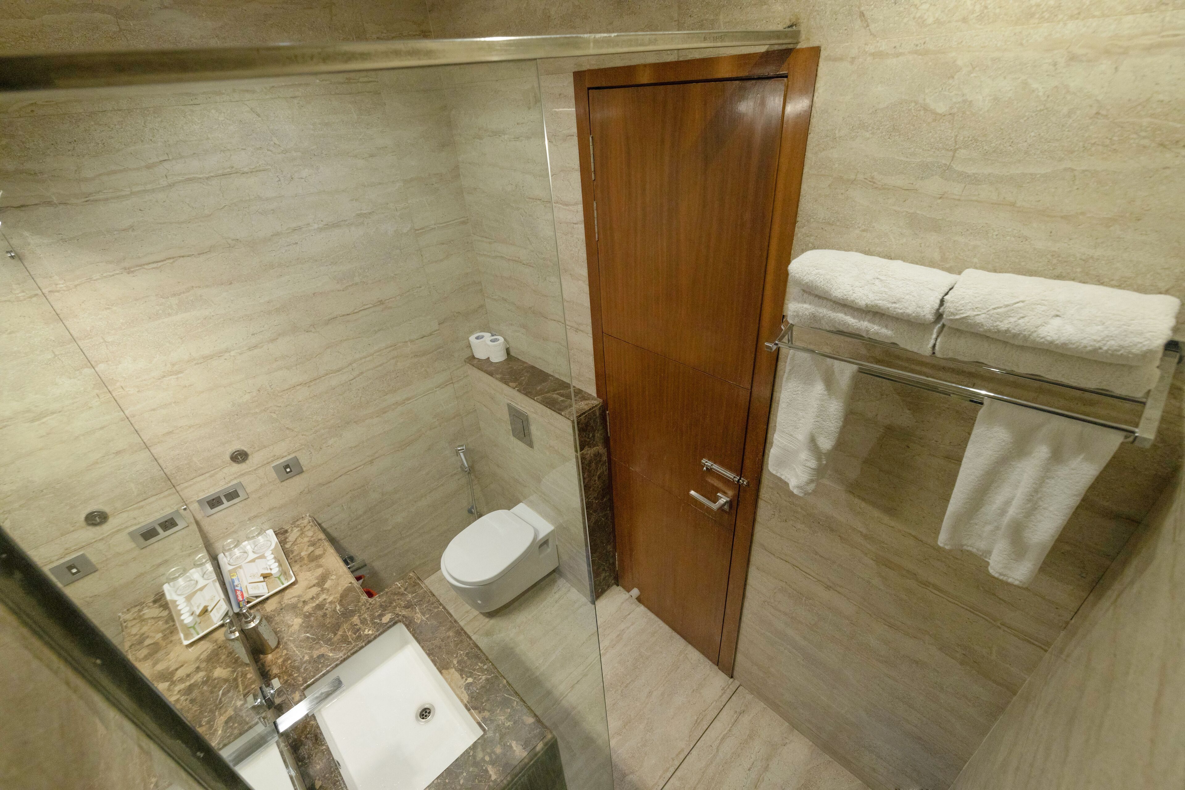 Un baño moderno con lavamanos, inodoro y ducha de mármol. Se observa una puerta de madera y un cerramiento de ducha de vidrio.