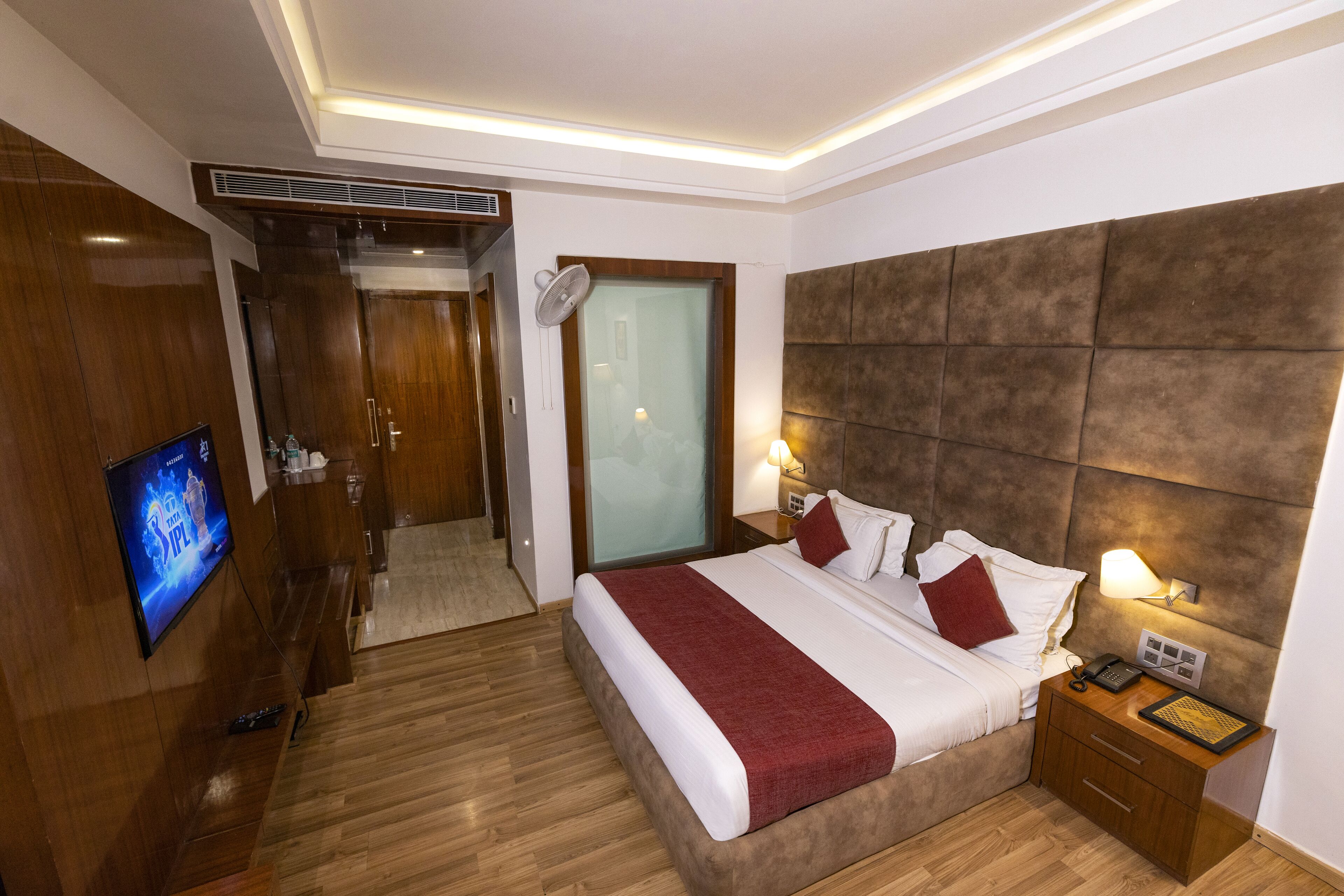 A hotel room with a large bed, wooden flooring, a TV on the wall, a bedside table with a lamp, and a wardrobe.