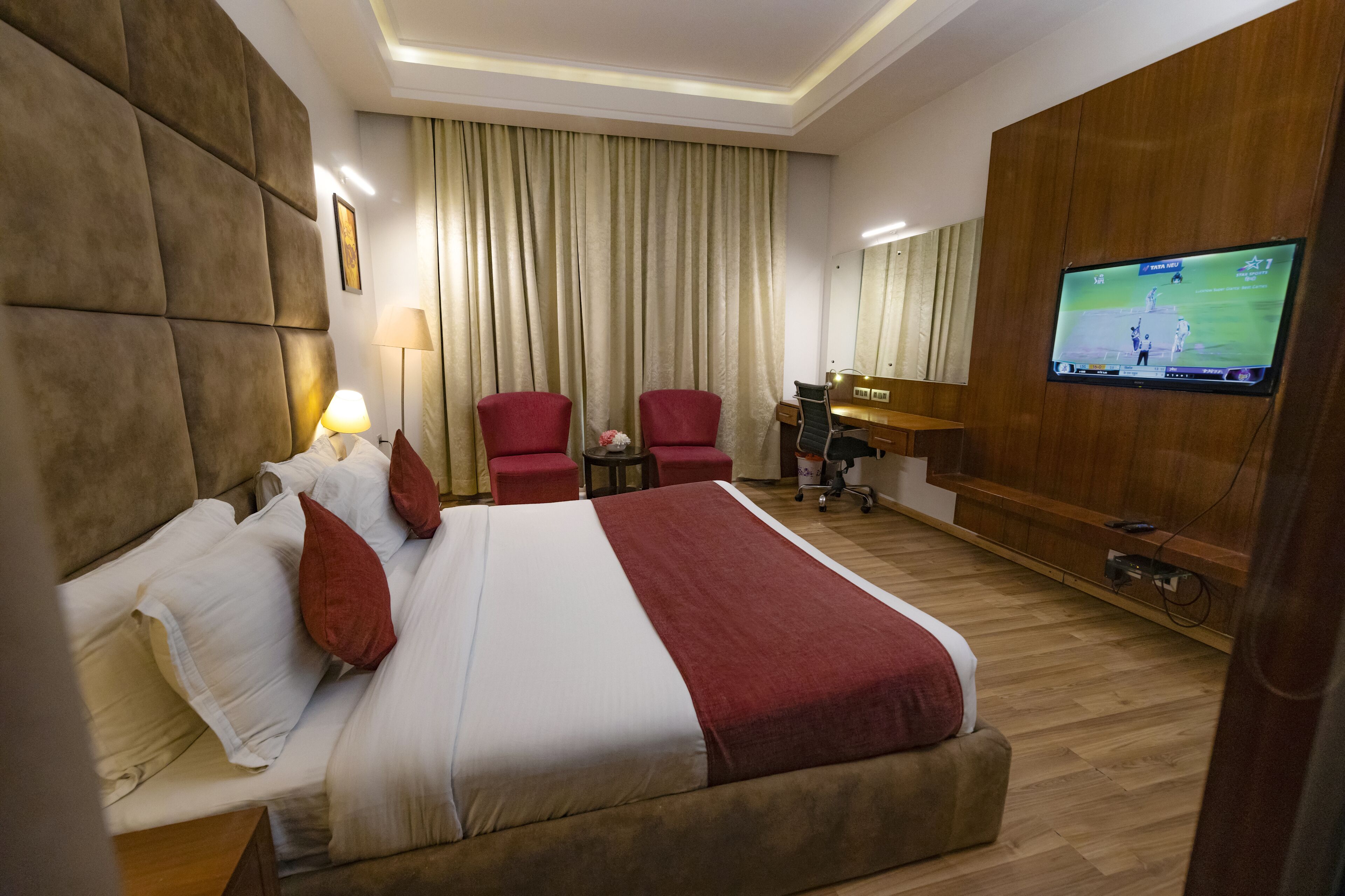 Habitación de hotel con una cama grande, dos sillas rojas, un escritorio, un televisor y suelo de madera.