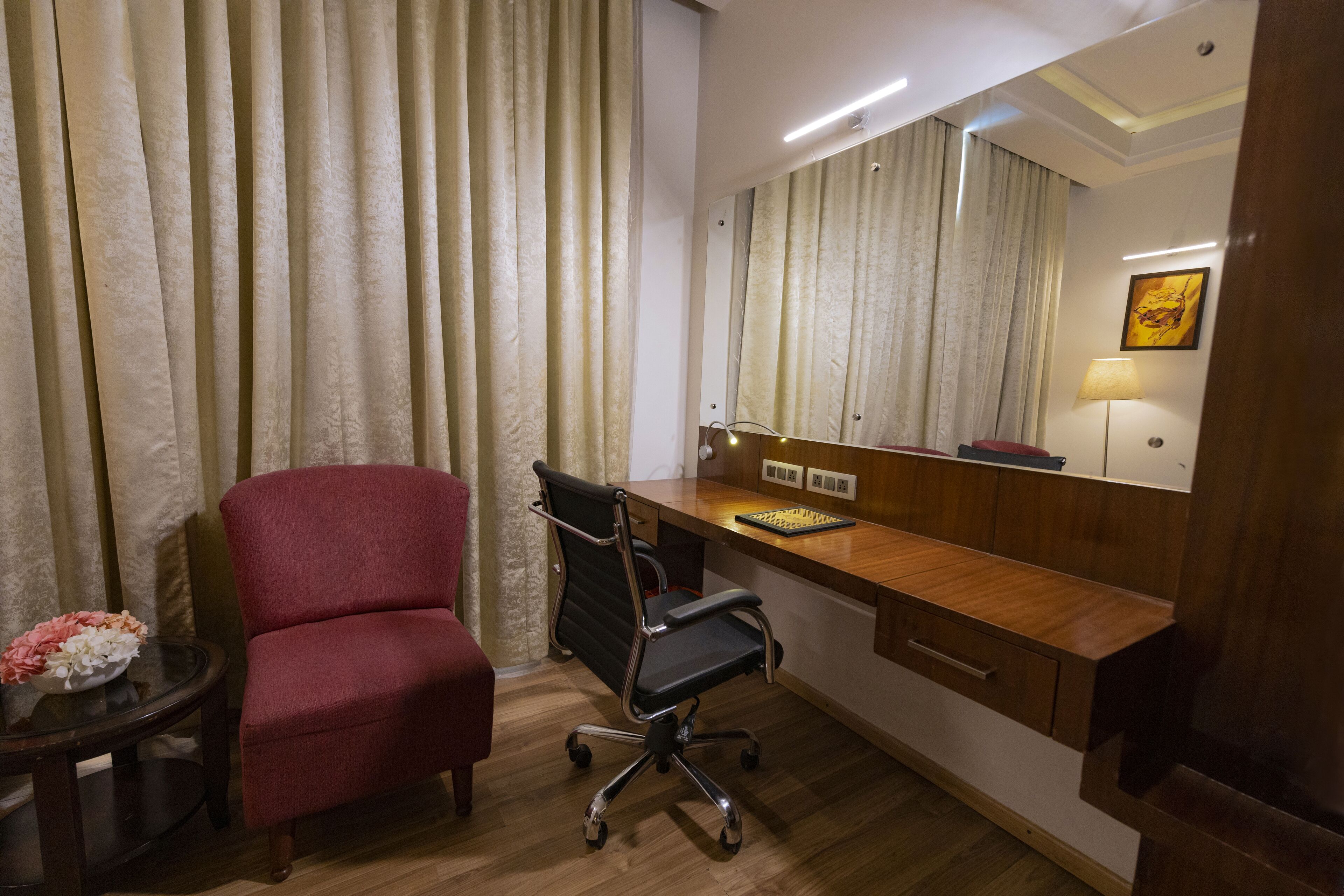 A hotel room with a wooden desk, a red chair, a black office chair, a small table with a flower vase, and a large mirror on the wall.