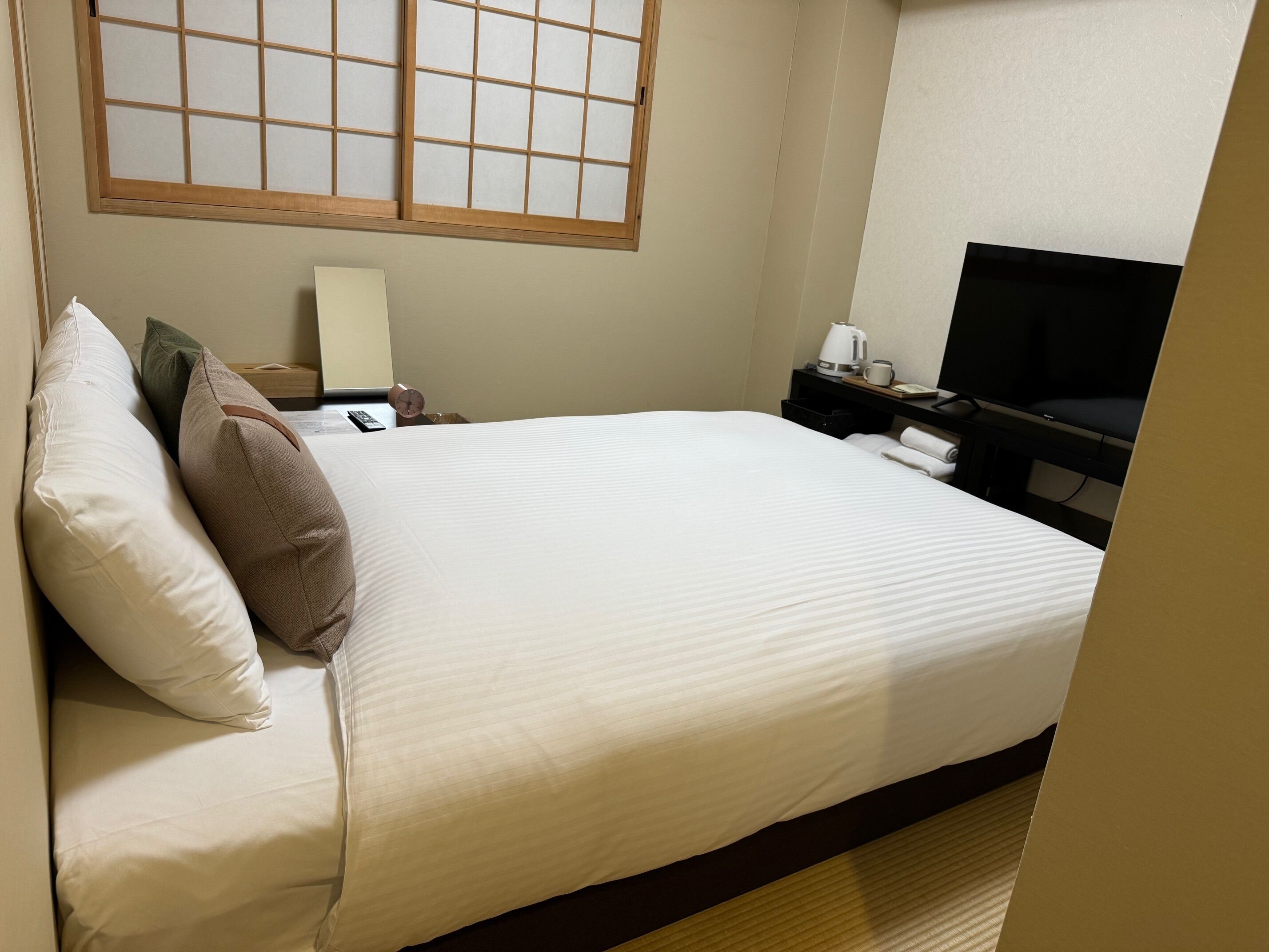 A hotel room with a bed, pillows, a bedside table with a lamp, a television, and a window with a grid pattern.