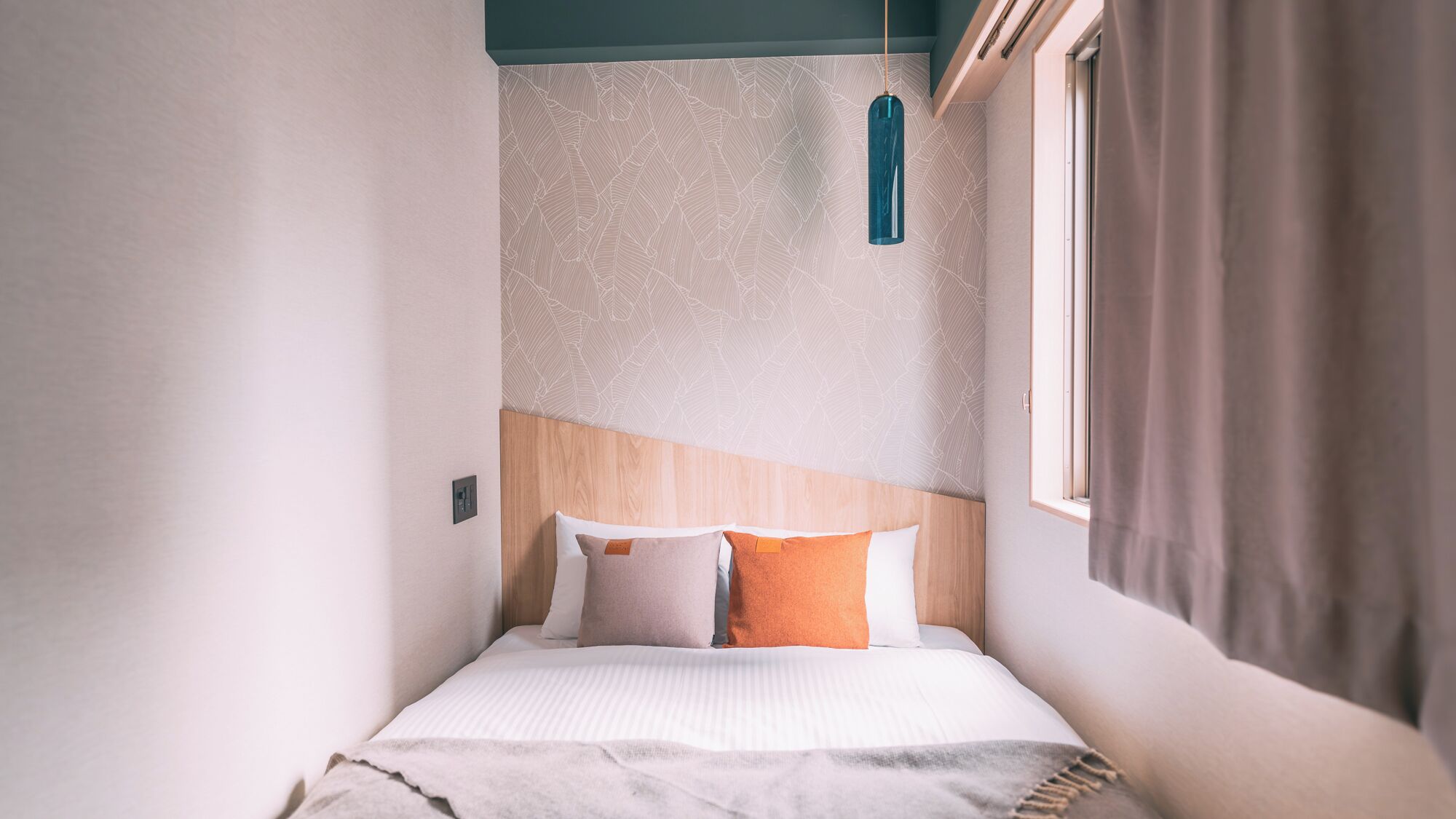 A narrow room with a bed, a hanging light fixture, and a window with curtains.