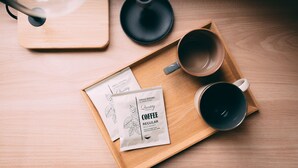 A wooden tray with two coffee packets, a black cup, and a black saucer on a wooden table. - R Hotel-The Atelier Shinsaibashi East (Osaka)