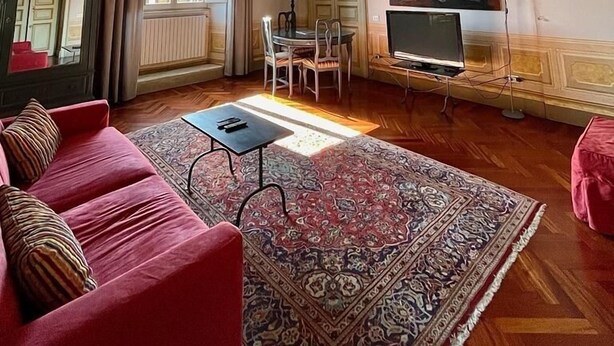 Ein geräumiges Wohnzimmer mit einem roten Sofa, einem gemusterten Teppich, einem Esstisch mit Stühlen, einem Fernseher und einem großen Fenster mit Vorhängen.