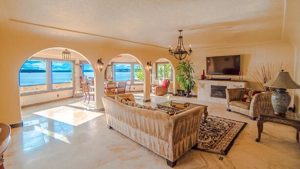 A spacious living room with a large sofa, a television, and a fireplace. There are arched windows providing a view of the sea.