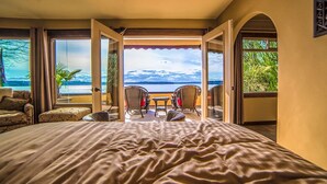 A bedroom with a large bed, a balcony with wicker chairs, and a view of the sea.