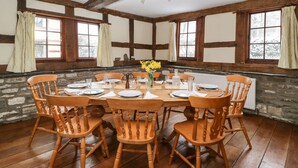 A dining room with a wooden table set for six, surrounded by six wooden chairs. The room features exposed wooden beams, large windows with curtains, and a stone wall. - The Old Rectory (Presteigne)