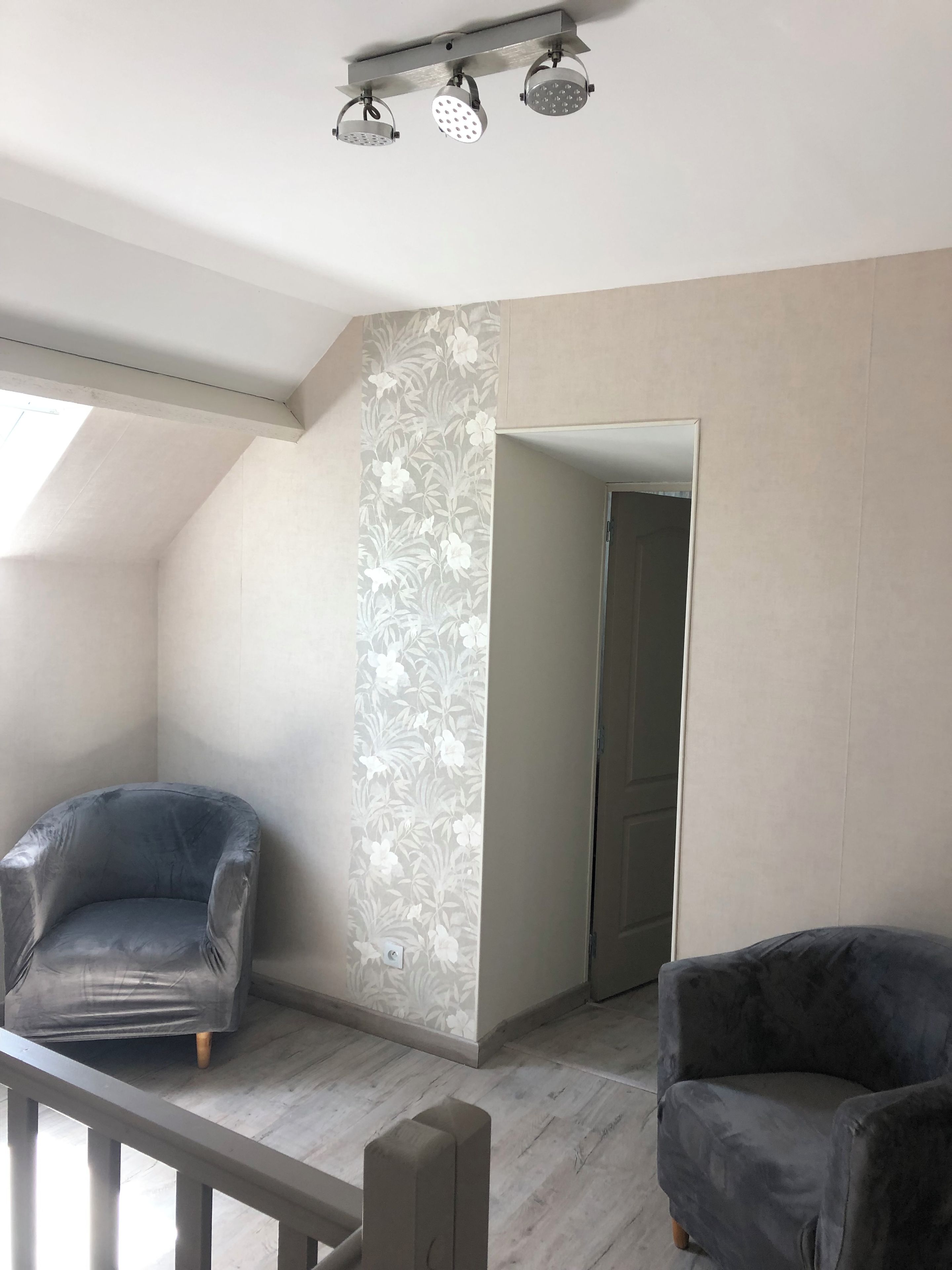 Une pièce avec un plafond en pente, un fauteuil gris, un mur à motifs et une porte donnant sur une autre pièce.