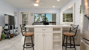 Una cocina moderna con una isla central, dos taburetes de bar, un ventilador de techo y un amplio ventanal con vistas a palmeras.