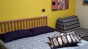 A room with a bed, a wall-mounted air conditioner, a hanging light fixture, a painting, and a small table with a lamp.
