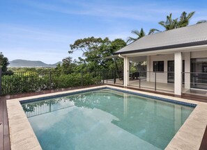 Relax by the serene pool surrounded by lush greenery and distant hills, with a covered seating area for shade and relaxation.