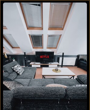 En moderne stue med et stort fladskærms TV, grå mønstrede sofaer, et sofabord og ovenlys.