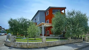 Ein modernes, zweistöckiges Gebäude mit roter und grauer Fassade, einem überdachten Außenbereich mit Sitzgelegenheiten und einem angelegten Garten mit Bäumen und einem Ziegelweg.