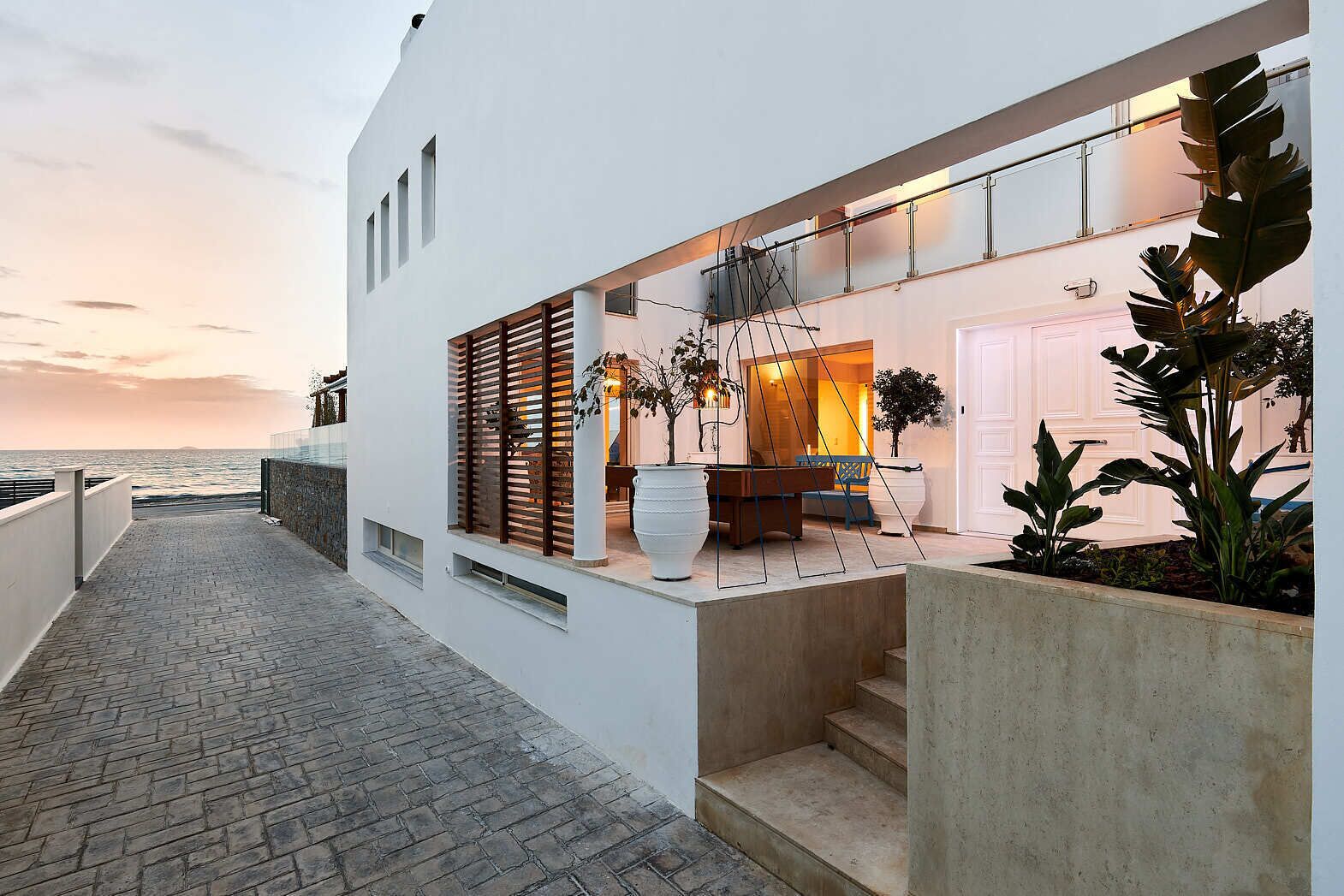 A modern building with a balcony overlooking the sea, featuring a dining area with a table and chairs, and large potted plants.