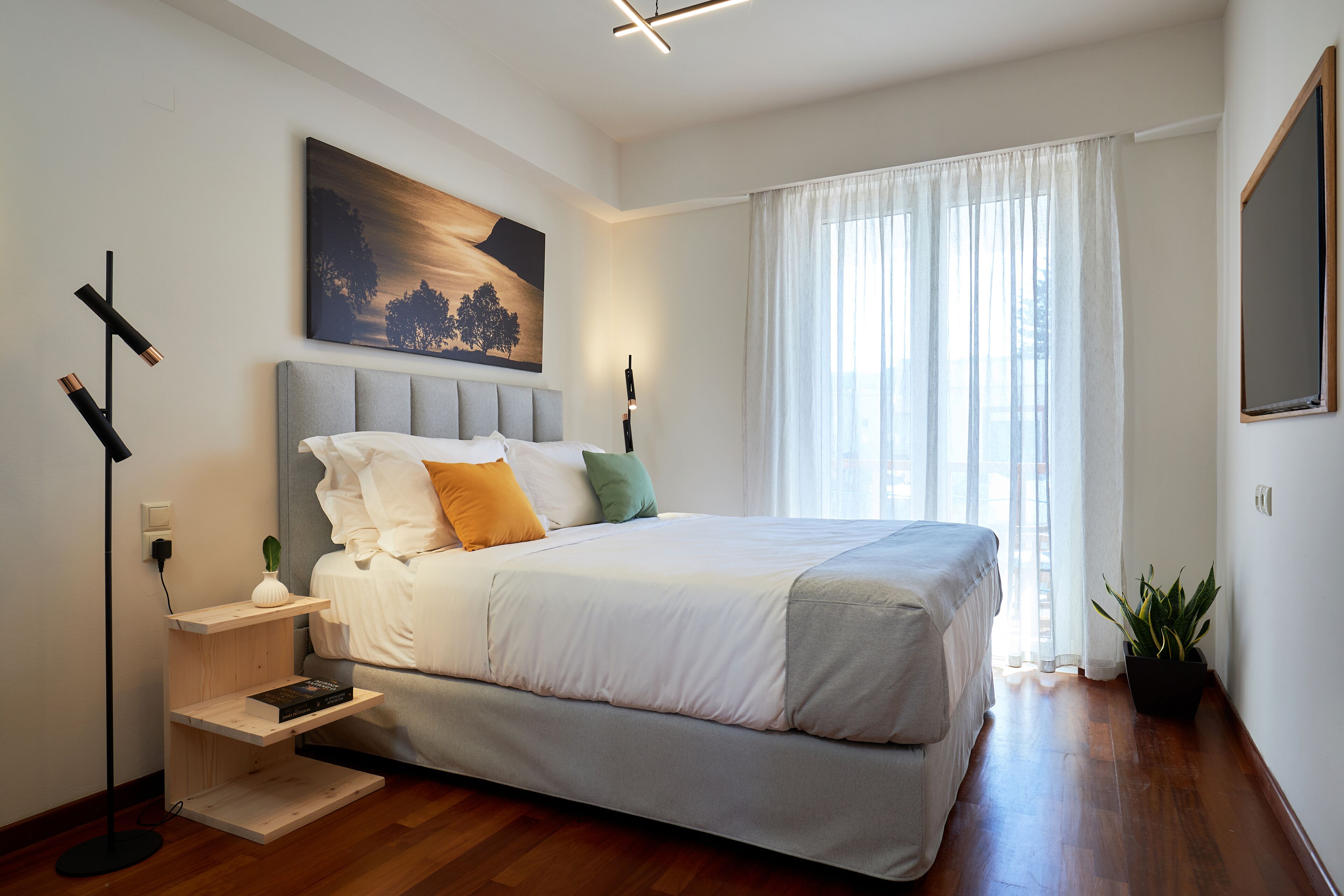 A modern bedroom with a large bed, a bedside table with a lamp, a painting on the wall, and a window with curtains.