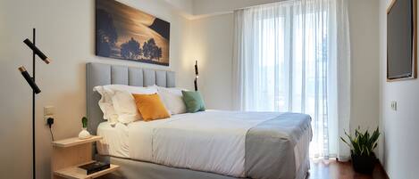 A modern bedroom with a large bed, a bedside table with a lamp, a painting on the wall, and a window with curtains.