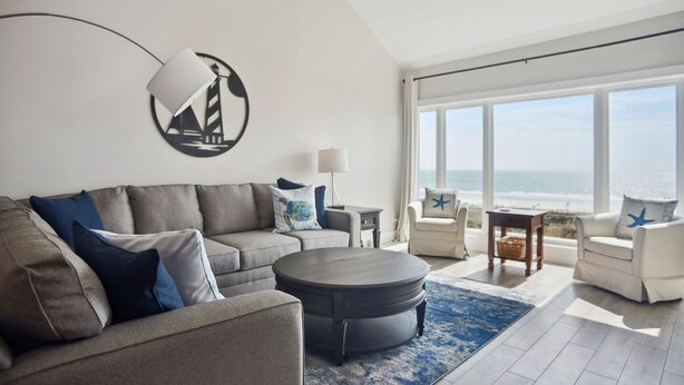A modern living room with a grey sofa, a round coffee table, and two armchairs. A large window offers a view of the ocean.