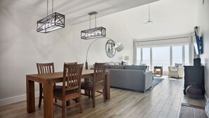 A spacious living room with a wooden dining table and chairs, a grey sofa, and a large window offering a view of the sea.