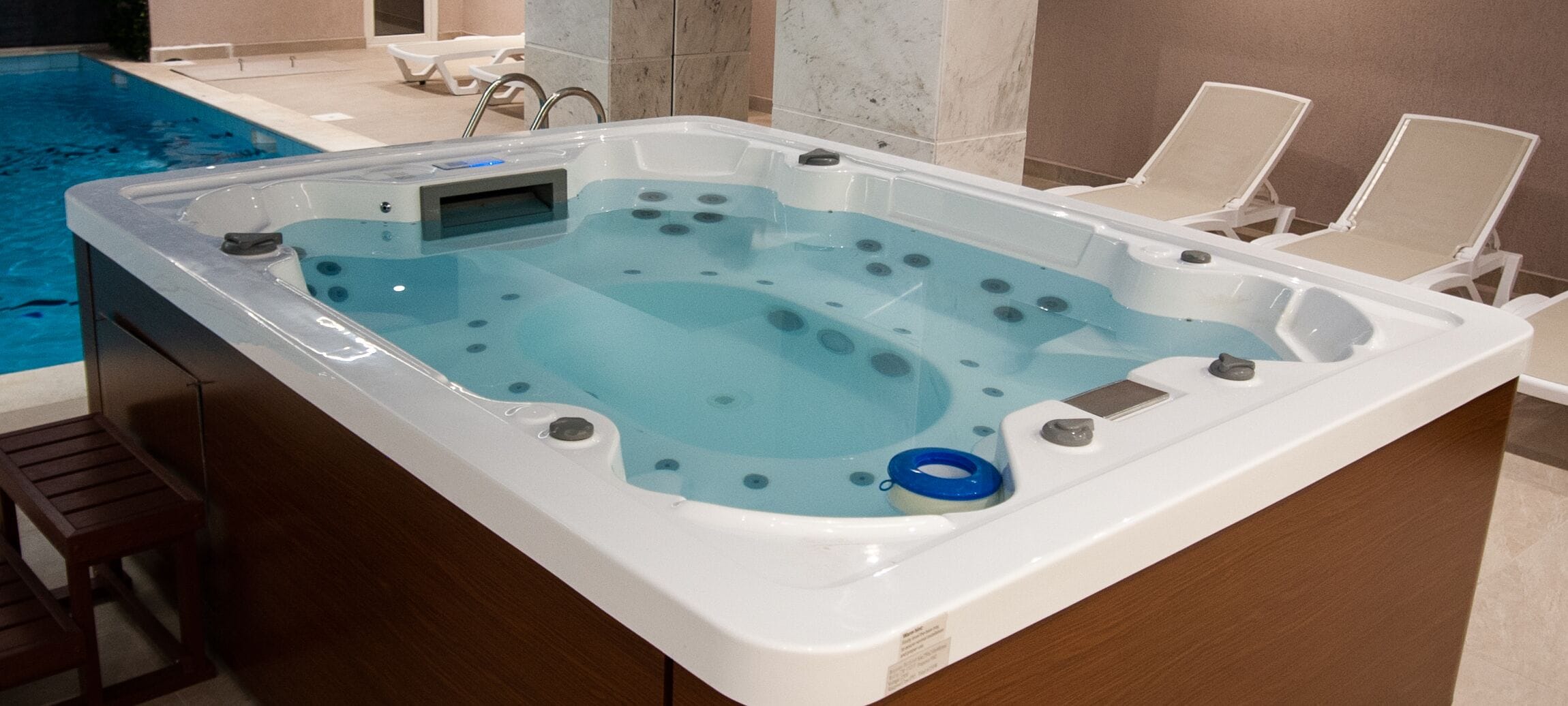 A hot tub with a control panel, two lounge chairs, and a pool in the background.