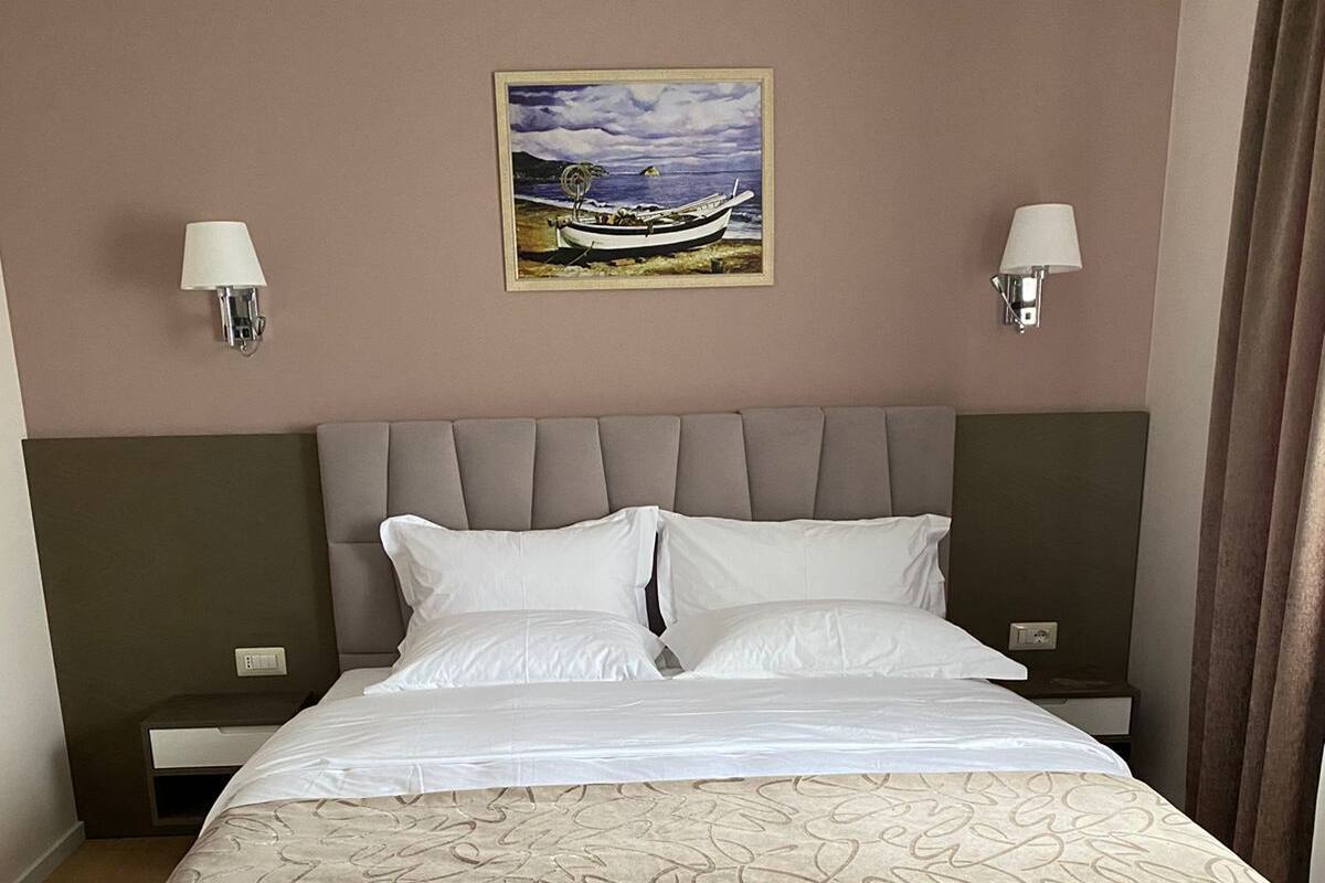 A neatly made bed with a beige and white bedspread, two bedside tables with lamps, a headboard, and a painting of a boat on the wall.