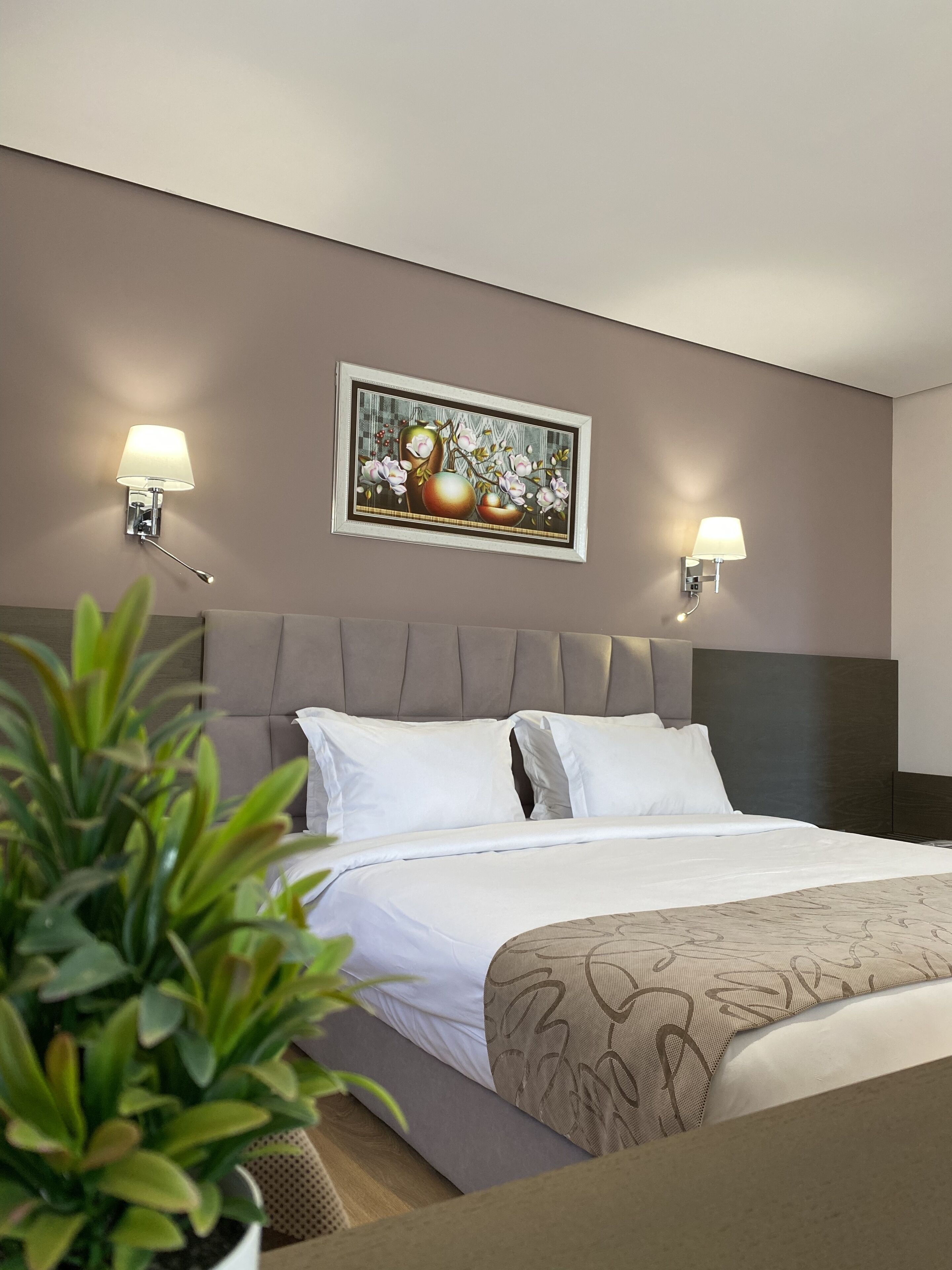 A hotel room with a large bed, two wall-mounted lamps, a framed artwork above the bed, and a potted plant in the foreground.