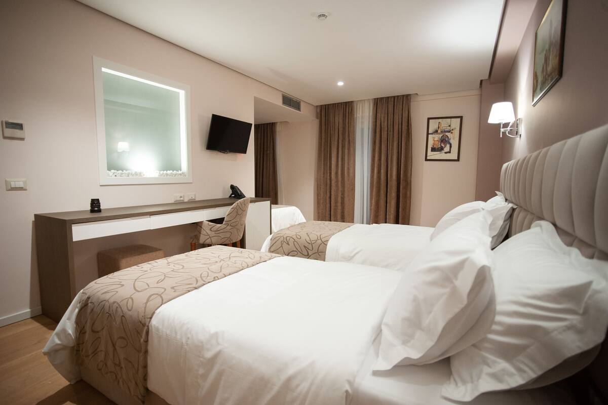 A hotel room with two beds, a desk, a flat-screen TV, and a large window with curtains.