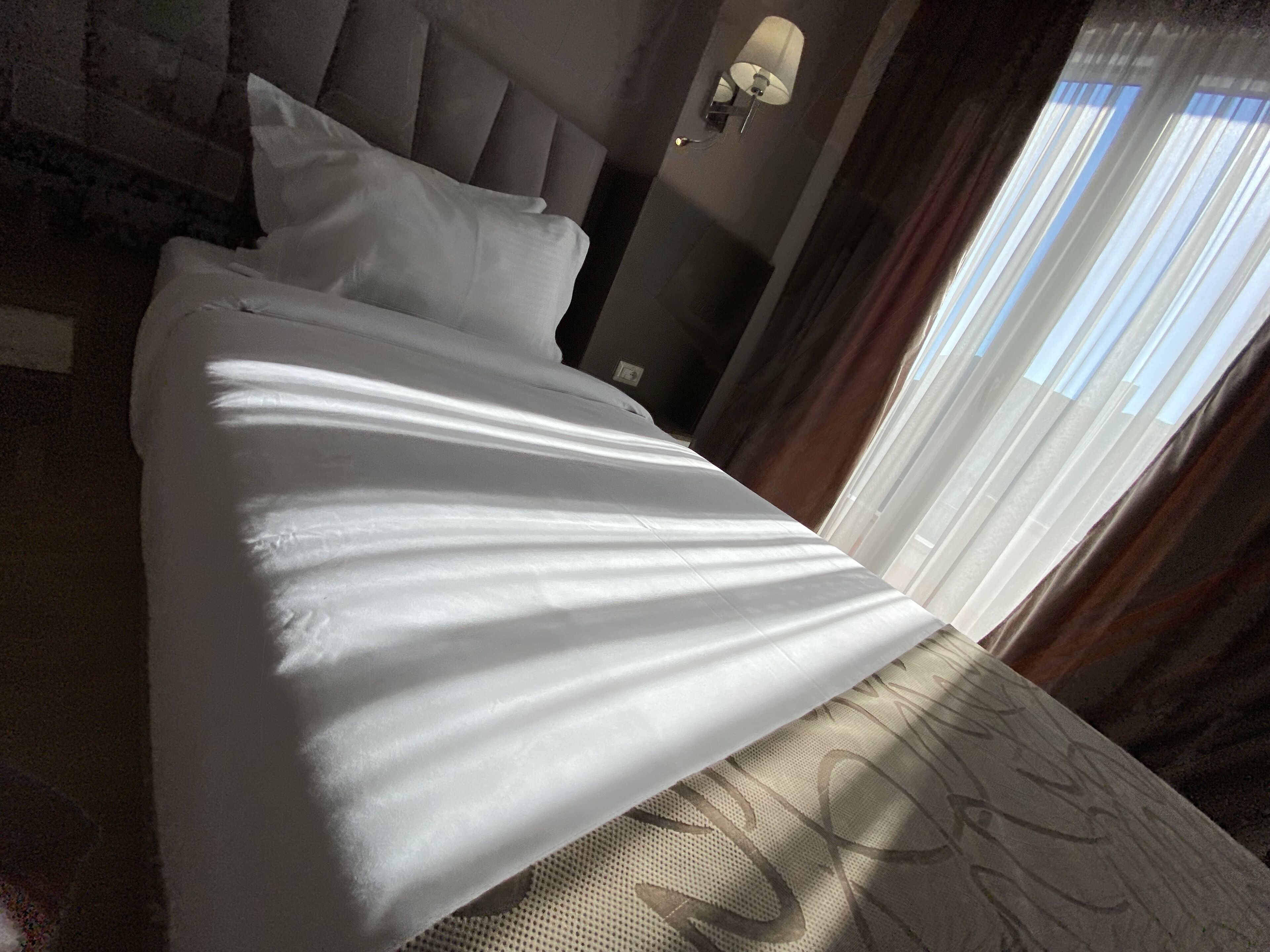 A neatly made bed with a headboard, a wall-mounted lamp, and a window with curtains.