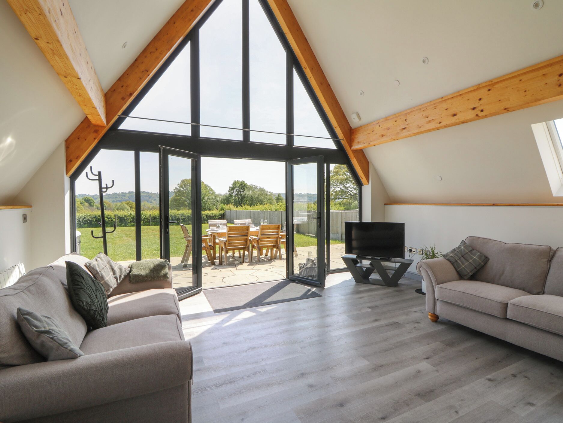 A spacious living area with a large sliding glass door leading to an outdoor patio. Inside, there is a sofa, a television on a stand, and a wooden ceiling with exposed beams.