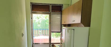 A narrow kitchen with a refrigerator, wooden cabinets, and a window with blinds.
