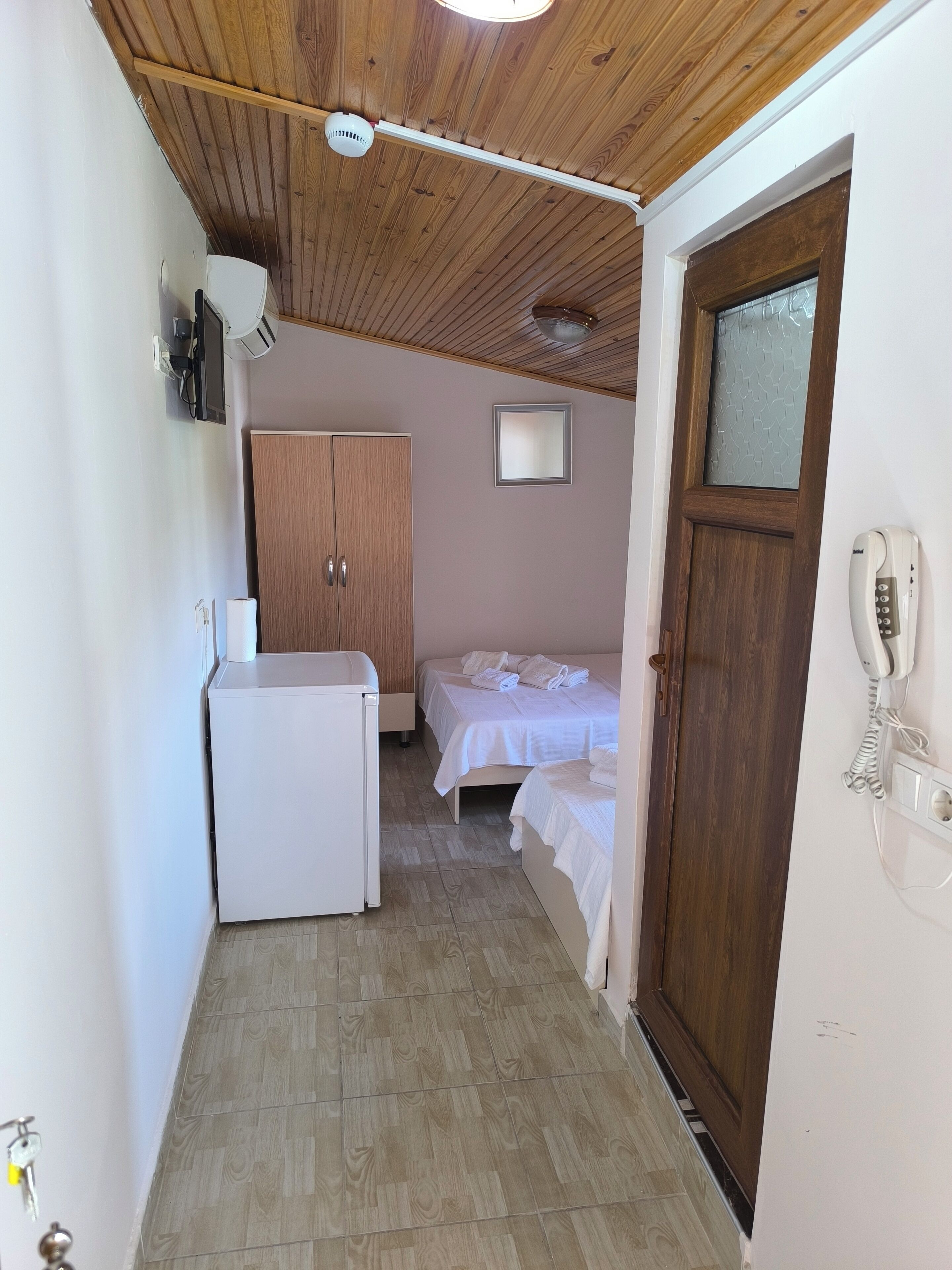 A narrow hallway with a wooden ceiling, a white refrigerator, a wooden wardrobe, and a bed with white bedding.