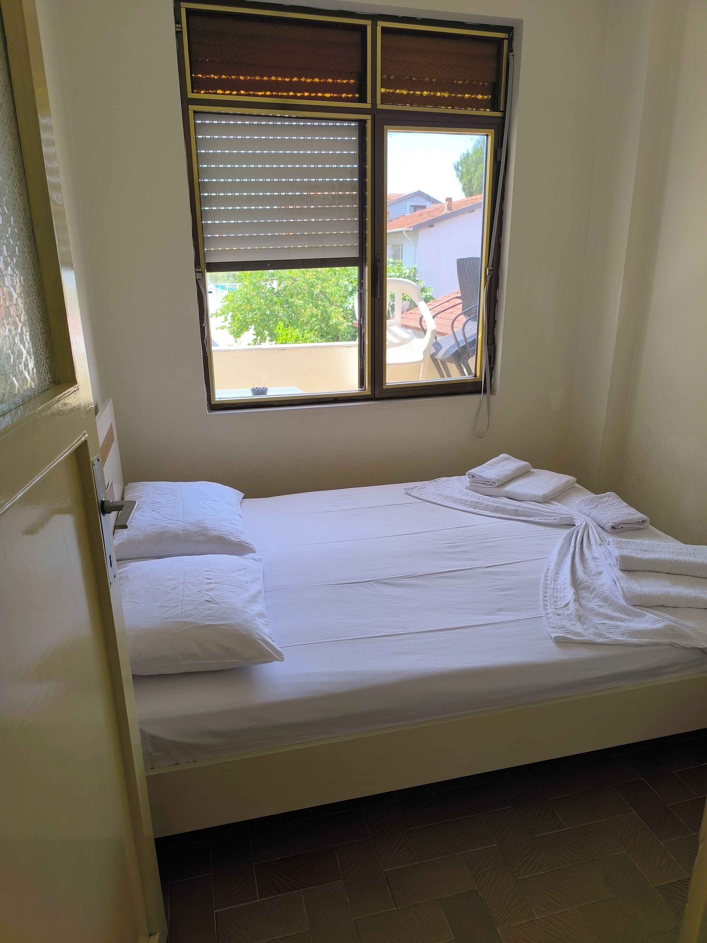 A single bed with white linens and towels, a window with blinds, and a view of a residential area outside.