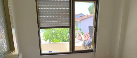 A single bed with white linens and towels, a window with blinds, and a view of a residential area outside.